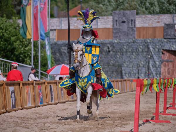 Ein Ritter in farbenfroher Rüstung reitet auf einem weißem Pferd in einer historischen Turnierkulisse.