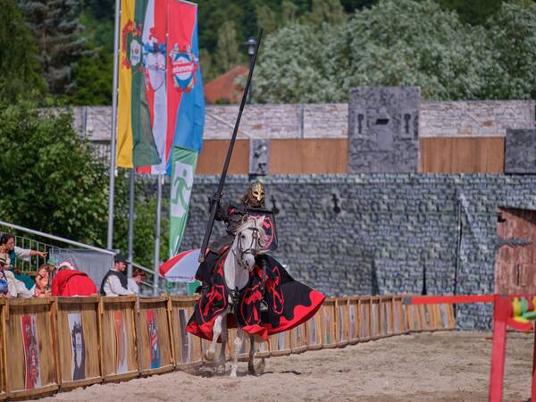 Ein Ritter in farbenfroher Rüstung reitet auf einem Pferd in einer historischen Turnierkulisse.