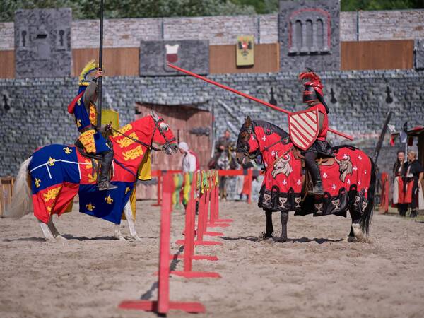 Zwei Ritter in farbenfroher Rüstung reiten auf Pferden in einer historischen Turnierkulisse.