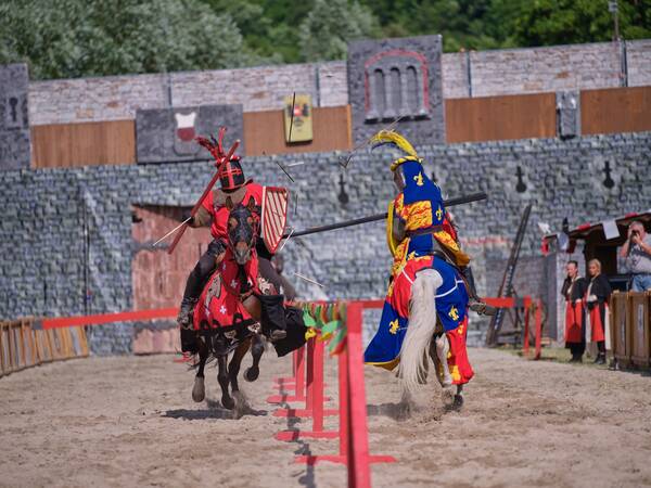 Zwei Ritter in farbenfrohen Rüstungen reiten während eines Turniers auf Pferden aufeinander zu, um zu kämpfen, wobei eine Burgmauer im Hintergrund zu sehen ist.