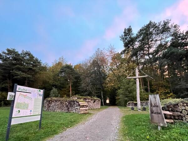 Waldweg mit Holzstapeln, einem großen Holzkreuz und einem Schild mit der Aufschrift 'Ruhe Wiese' am Eingang zum RuheWald.