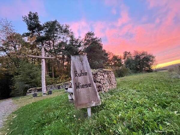 Holzschild mit der Aufschrift 'Ruhe Wiese' vor einem Holzstapel und einem großen Holzkreuz bei Sonnenuntergang.