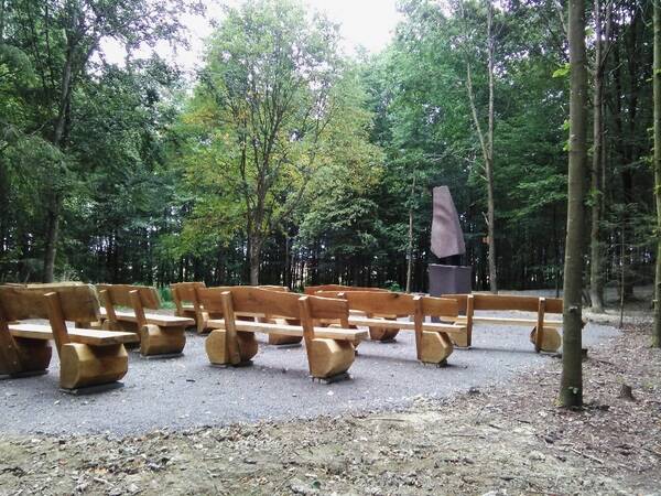 Holzbänke in einem Wald, angeordnet vor einer Steinskulptur auf einem Kiesplatz.