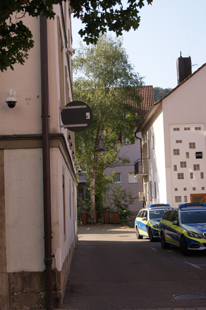 Enger Durchgang zwischen Gebäuden mit zwei geparkten Polizeiautos und Bäumen im Hintergrund.