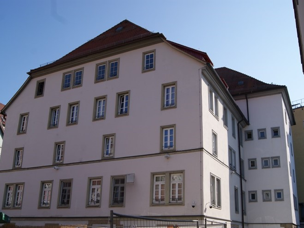Mehrstöckiges weißes Gebäude mit roten Dachziegeln in einer städtischen Umgebung bei klarem Himmel.