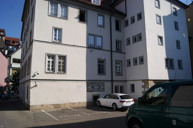 Weißes mehrstöckiges Gebäude mit zahlreichen Fenstern und zwei geparkten Autos auf einem Parkplatz davor.