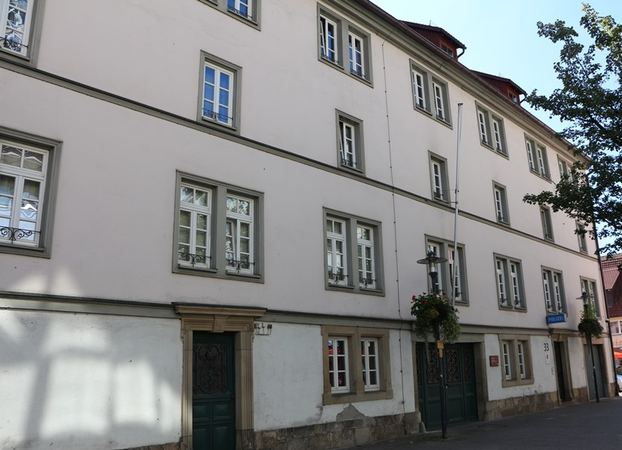 Das Bild zeigt ein großes, mehrstöckiges Gebäude mit weißen Wänden, zahlreichen Fenstern und einem Eingang mit grüner Tür in einer städtischen Umgebung.