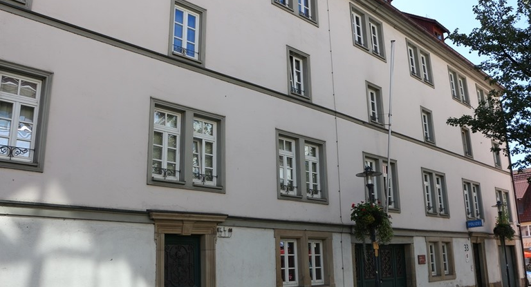 Das Bild zeigt ein großes, mehrstöckiges Gebäude mit weißen Wänden, zahlreichen Fenstern und einem Eingang mit grüner Tür in einer städtischen Umgebung.