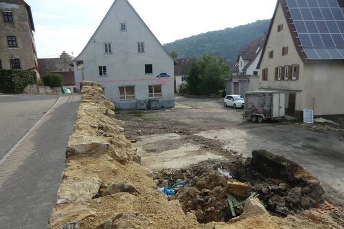 Blick auf eine Baustelle mit freigelegtem Erdreich und einer niedrigen Steinmauer, umgeben von Wohnhäusern in einem Dorf