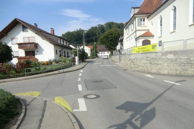 Leere Straße in einem Dorf mit weißen Häusern, Steinmauer und grünem Hügel im Hintergrund.