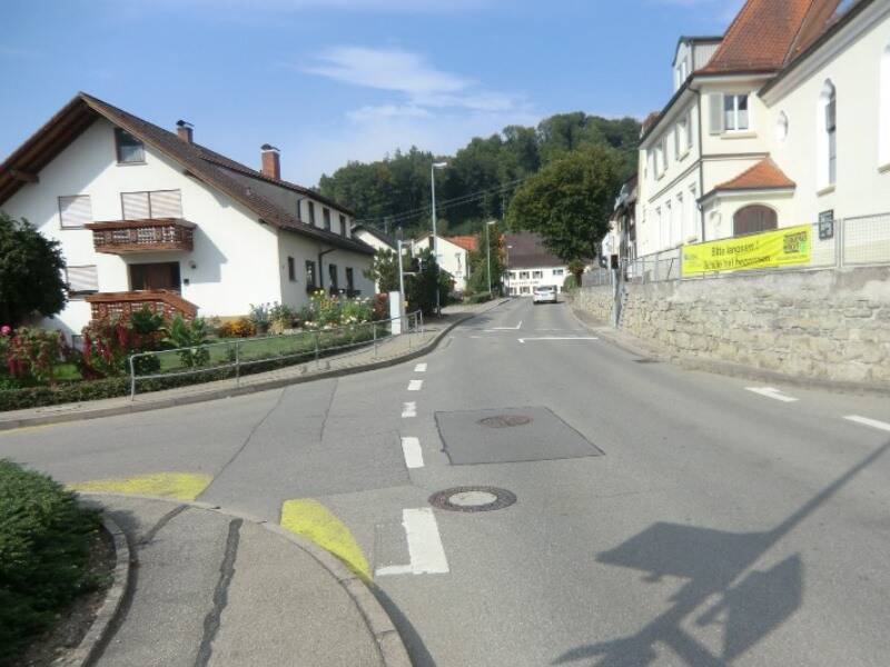 Leere Straße in einem Dorf mit weißen Häusern, Steinmauer und grünem Hügel im Hintergrund.