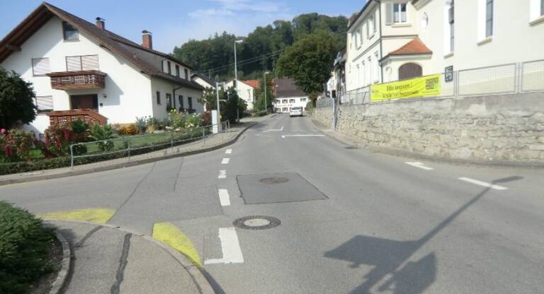 Leere Straße in einem Dorf mit weißen Häusern, Steinmauer und grünem Hügel im Hintergrund.