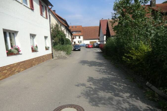 Dorfstraße mit weißen Häusern, blühenden Fensterkästen und parkenden Autos unter blauem Himmel.
