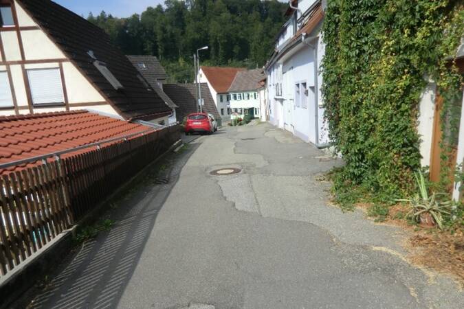 Enge Straße in einem Dorf mit Fachwerkhäusern, einem roten Auto und bewaldeten Hügeln im Hintergrund.