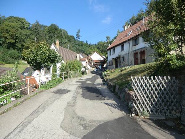 Schmale Straße in einem Dorf mit alten Häusern und bewaldetem Hügel im Hintergrund bei klarem Himmel.
