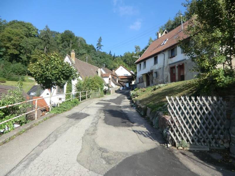 Schmale Straße in einem Dorf mit alten Häusern und bewaldetem Hügel im Hintergrund bei klarem Himmel.