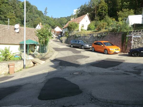Dorfstraße mit geparkten Autos, Häusern und bewaldeten Hügeln im Hintergrund bei sonnigem Wetter.