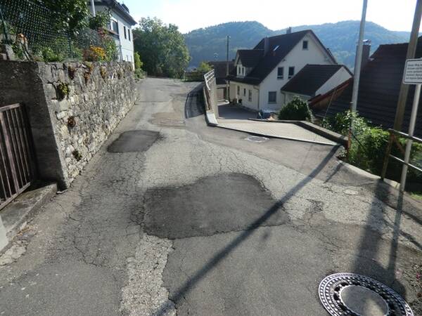 Schmale Straße mit Flickstellen im Asphalt, links eine Steinmauer, rechts Häuser und Hügel im Hintergrund.