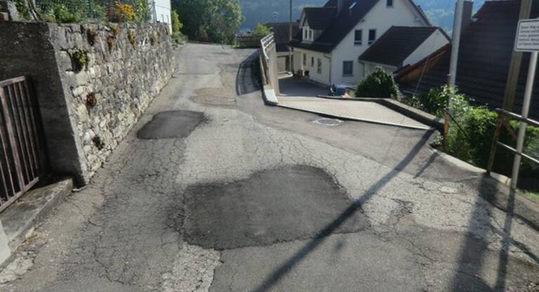 Schmale Straße mit Flickstellen im Asphalt, links eine Steinmauer, rechts Häuser und Hügel im Hintergrund.