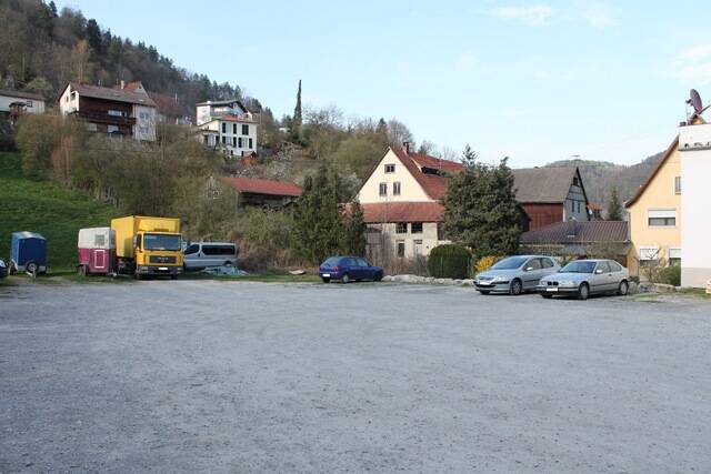 Parkplatz mit mehreren geparkten Autos und einem gelben Lastwagen vor Häusern und bewaldeten Hügeln.
