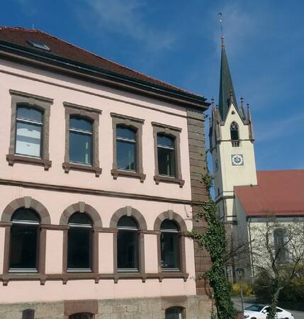 Fassade eines zweistöckigen Gebäudes mit sechs Fenstern und einem Kirchturm mit Uhr und spitzem Dach im Hintergrund.