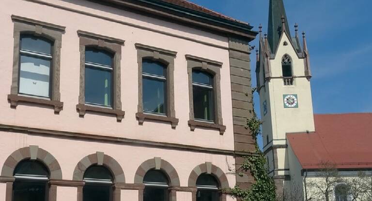 Fassade eines zweistöckigen Gebäudes mit sechs Fenstern und einem Kirchturm mit Uhr und spitzem Dach im Hintergrund.