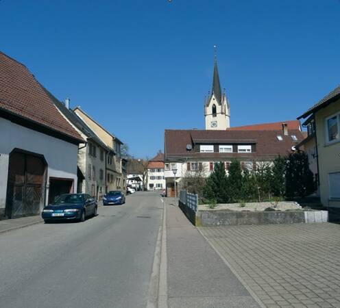 Straße in einem Dorf mit geparkten Autos, Wohnhäusern und einer Kirche mit hohem Turm im Hintergrund.