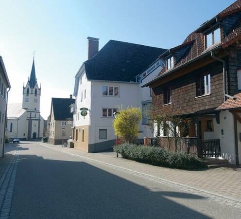 Straße in einem Dorf mit traditionellen Häusern und einer Kirche mit hohem Turm im Hintergrund.