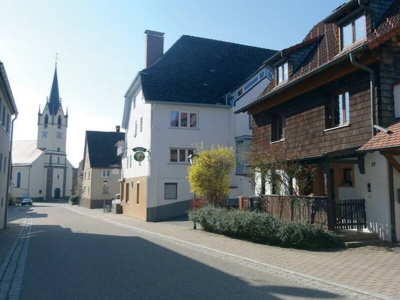 Straße in einem Dorf mit traditionellen Häusern und einer Kirche mit hohem Turm im Hintergrund.
