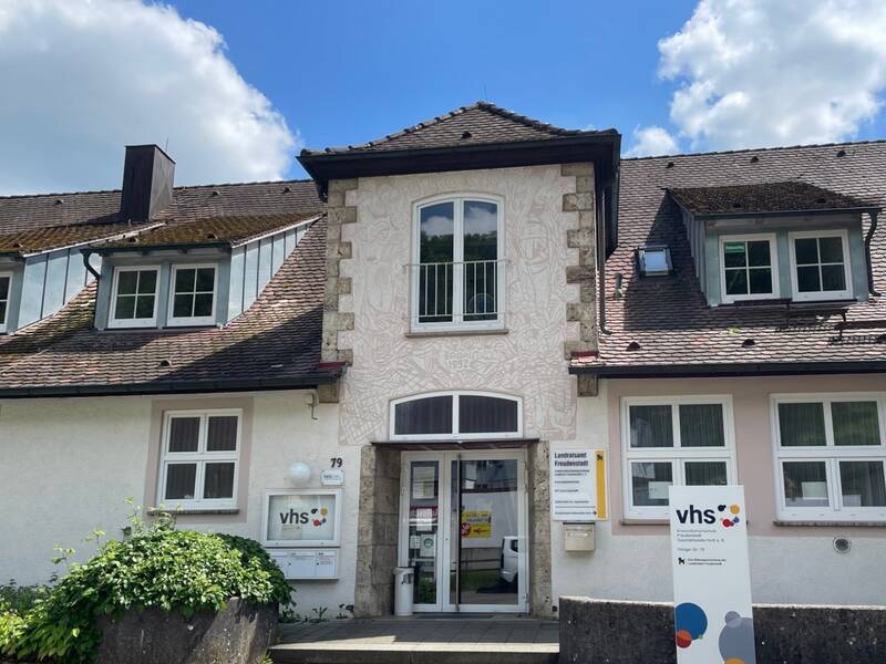 Eingang eines Gebäudes mit Schildern der Volkshochschule (vhs) und einem Schild mit Öffnungszeiten.
