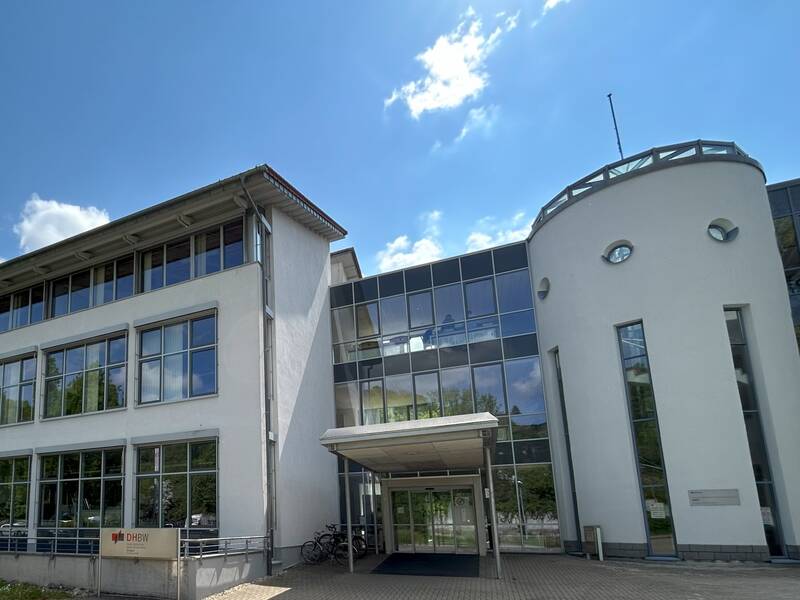 Modernes Bürogebäude mit großen Fenstern und rundem Turm unter blauem Himmel mit Wolken.
