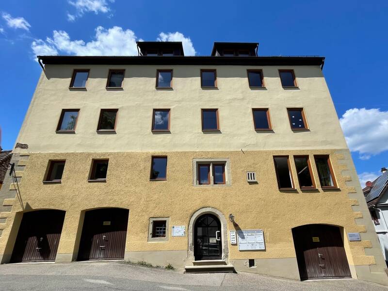Mehrstöckiges Gebäude mit gelblich-beiger Fassade, mehreren Fenstern und drei Torbögen im Erdgeschoss unter blauem Himmel.