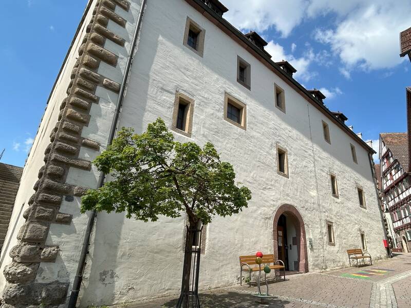 Altes weißes Gebäude mit kleinen Fenstern und einem Torbogen, davor ein Baum und zwei Bänke auf gepflastertem Platz.