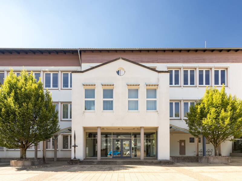 Ein dreistöckiges Gebäude mit vielen Fenstern und einem Eingangsbereich, flankiert von zwei großen Bäumen, ist vor einem klaren blauen Himmel zu sehen.