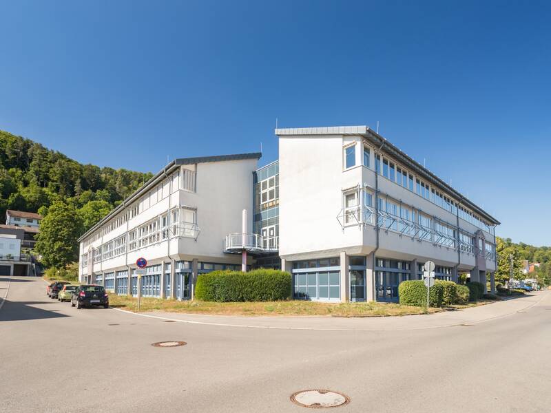 Ein modernes Gebäude mit weißen Wänden und großen Fenstern besitzt eine überdachte Verbindung zu einem weiteren Gebäudeteil und befindet sich in einem gepflasterten Außenbereich.