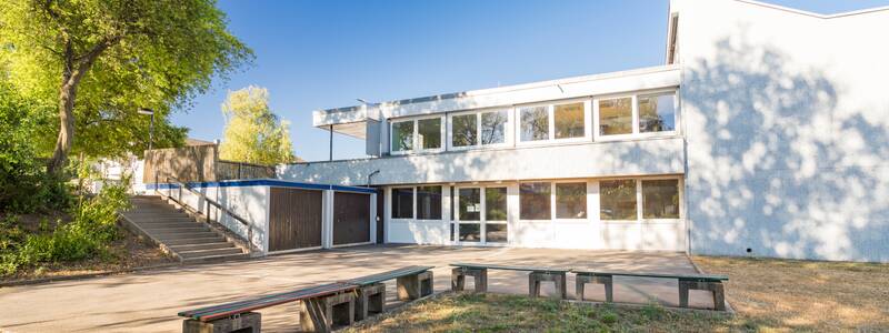 Ein zweistöckiges, modernes Gebäude mit großen Fenstern steht an einem sonnigen Tag neben einem kleinen Hof mit Sitzgelegenheiten und einer Treppe, die von grünem Laub flankiert ist.