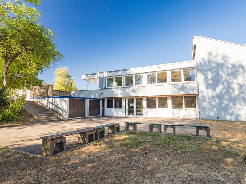 Ein zweistöckiges, modernes Gebäude mit großen Fenstern steht an einem sonnigen Tag neben einem kleinen Hof mit Sitzgelegenheiten und einer Treppe, die von grünem Laub flankiert ist.