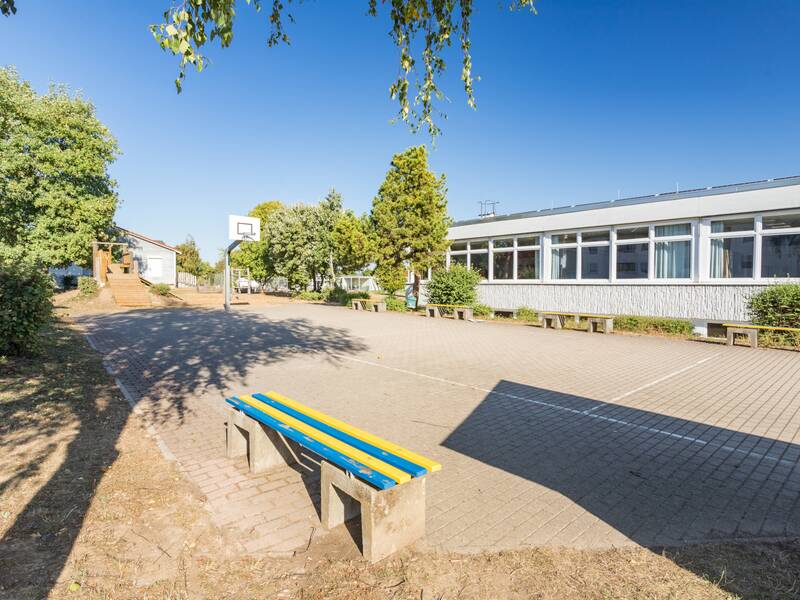 Ein leerer Schulhof mit einem Basketballkorb, einem blau-gelben Tisch und einem langen Gebäude mit großen Fenstern an einem sonnigen Tag.