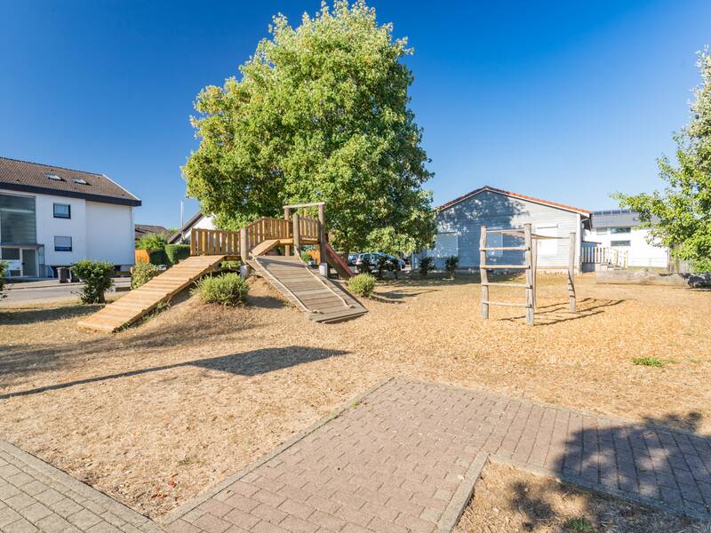 Ein Spielplatz mit Holzklettergerüst und Rutschen steht zwischen grünen Bäumen neben Wohnhäusern auf einem sonnigen Platz.