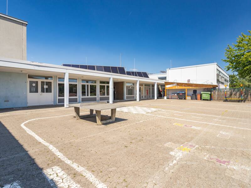 Das Bild zeigt den Schulhof mit einer Tischtennisplatte unter einem blauen Himmel.