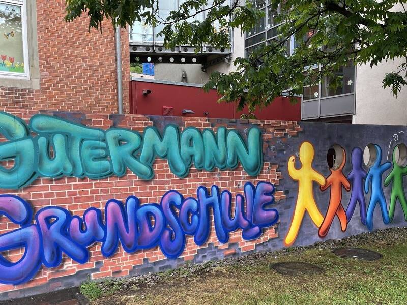 Eine bunte Graffiti-Wand zeigt die Wörter "Gutermann Grundschule" und stilisierte, farbenfrohe Figuren vor einer Ziegelsteinmauer.