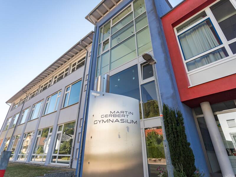 Ein modernes Schulgebäude mit großen Fenstern und einem Schild mit der Aufschrift "Martin Gerbert Gymnasium" ist zu sehen.