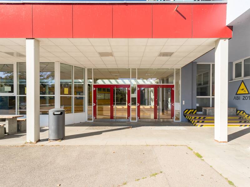 Schuleingang mit vielen Fenstern und zwei Glastüren und einem überdachten Bereich.