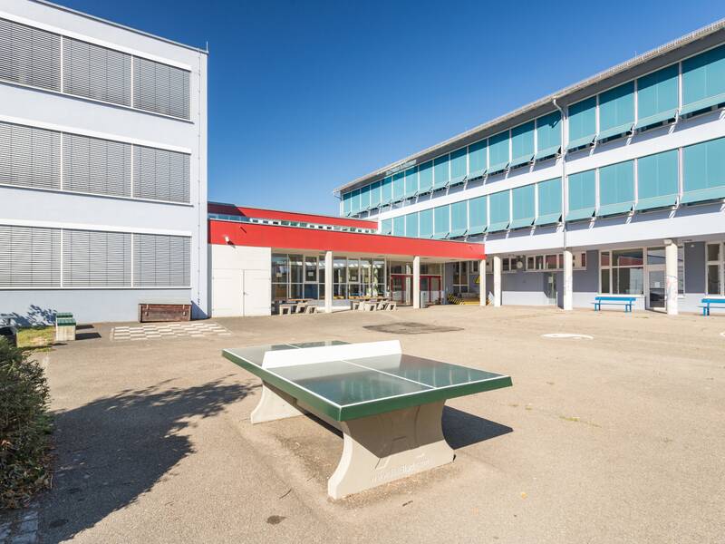 Das Bild zeigt den Schulhof mit einer Tischtennisplatte und einem eingang unter einem blauen Himmel.