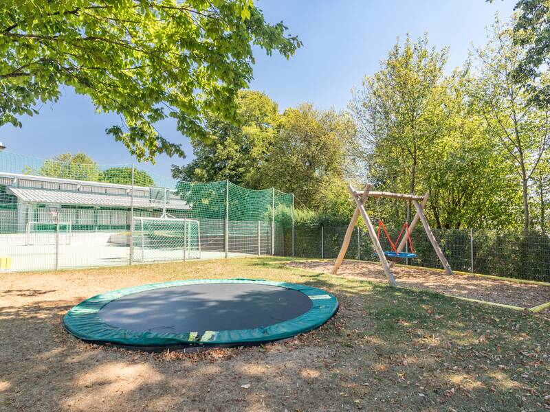 Ein Spielplatz mit einem Trampolin im Boden und einer Doppelschaukel, umgeben von Bäumen und einem Zaun.