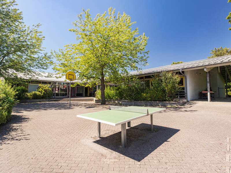 Das Bild zeigt den Schulhof mit einer Tischtennisplatte und einem Basketballkorb unter einem blauen Himmel.