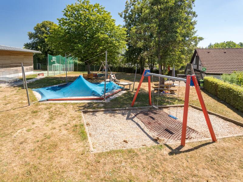 Ein Spielplatz mit einem Trampolin im Boden und einer Doppelschaukel, umgeben von Bäumen und einem Zaun.