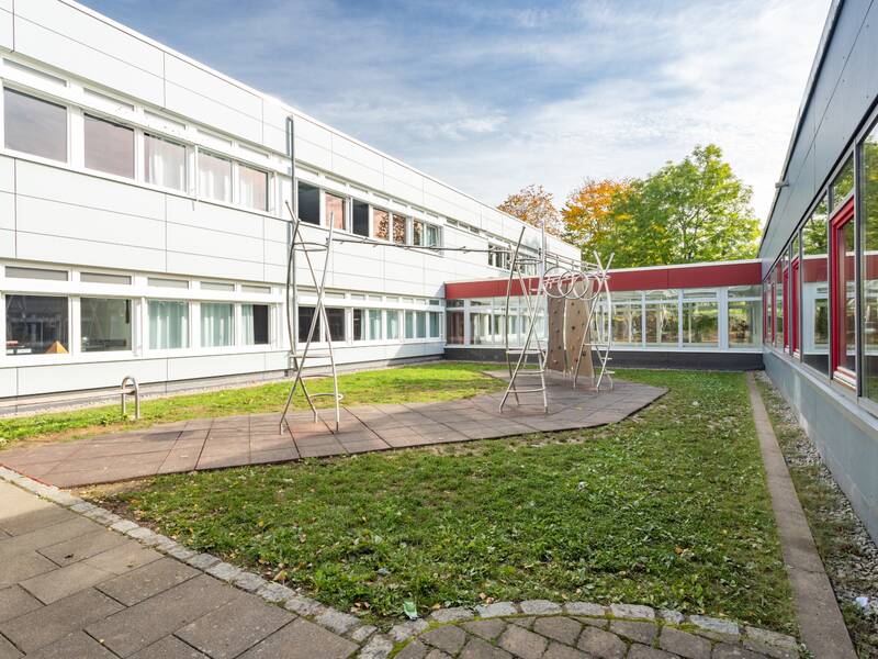 Ein Schulhof mit Klettergerüst, umgeben von einem weißen, einstöckigen Schulgebäude mit vielen Fenstern und grüner Rasenfläche.