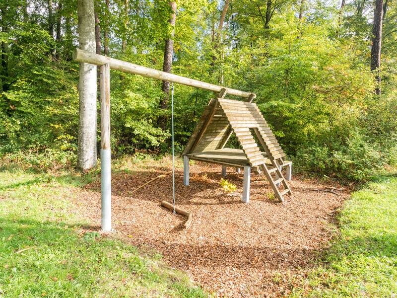 Eine Holzwippe und ein Klettergerüst in Form eines Tipis stehen auf einem mit Holzspänen bedeckten Spielplatz, umgeben von üppigem Grün.