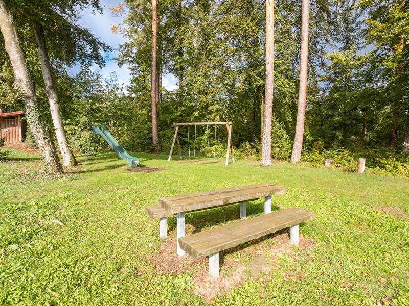 Blich auf grüne Rutsche und einer Doppelschaukel im Vordergrund Picknick Tisch mit zwei Bänken am Waldrand 